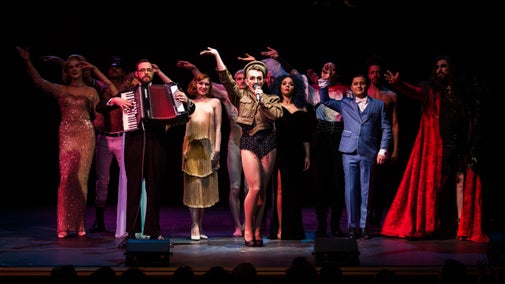 Image of Queerstory performers on stage, one holding a mic and several with one arm in the air, mid-dance move. Performers wear a variety of flamboyant costumes.